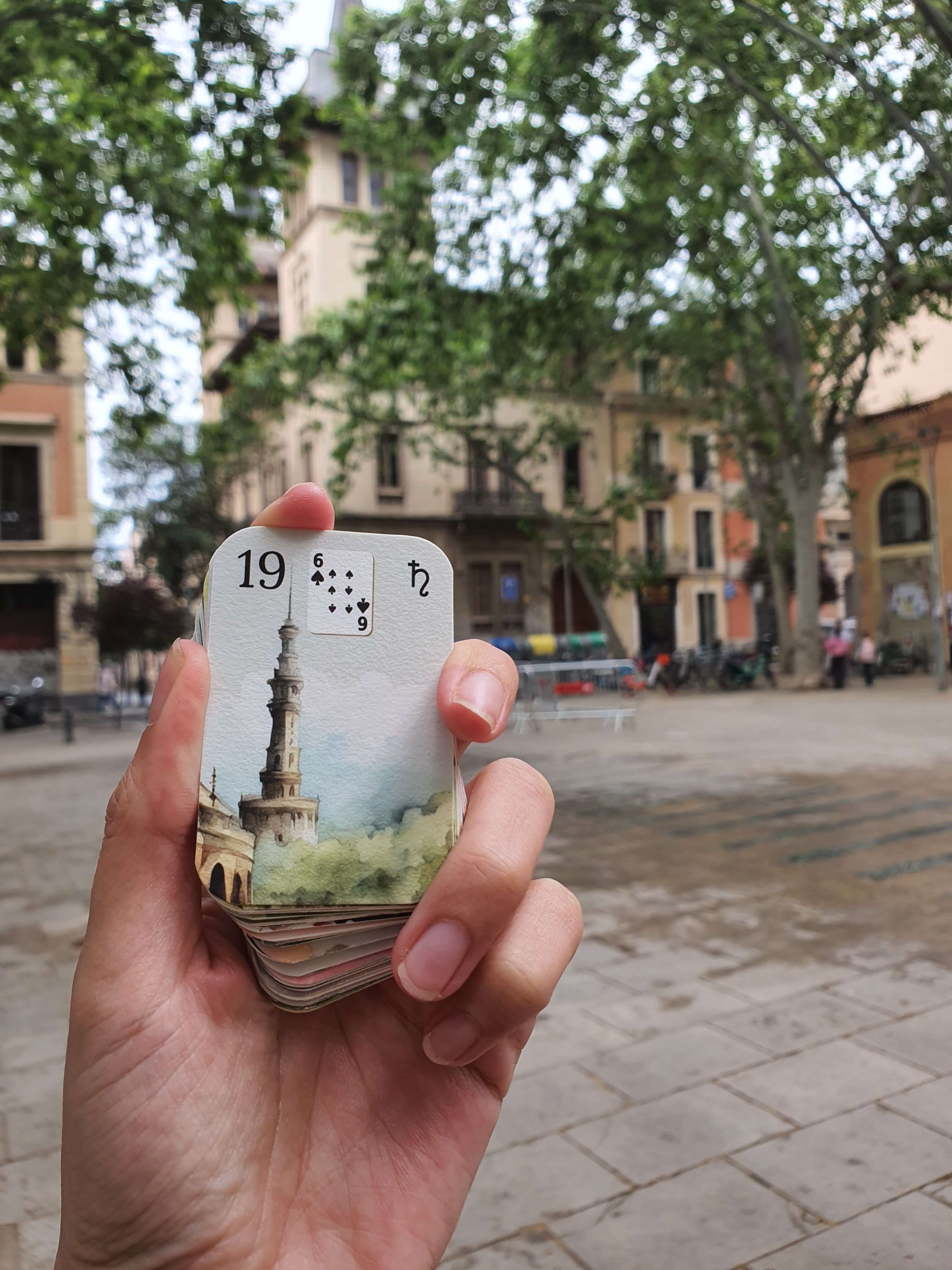 Petit Lenormand watercolor mini deck held in hand in front of a tree-lined square